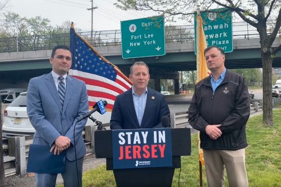 Rep. Josh Gottheimer, in a jacket and shirt, stands behind a lectern with a sign that says "Stay in New Jersey" beside a highway